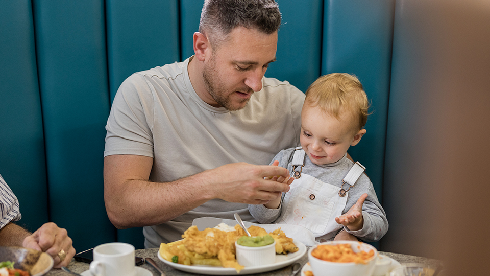Father feeding a toddler seated at a table in a booth.