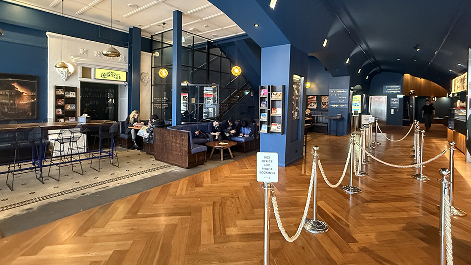 Interior photo of Classic Cinemas in Elsternwick Village. White ropes linking bollards in the foreground for queuing. Tables and booths for comfortable waiting next to an alternative entrance within the cinema foyer that leads to Copycat Restaurant and Bar.