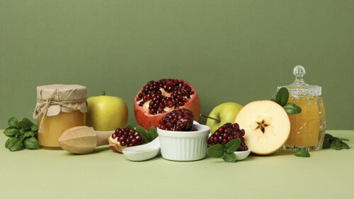 Fresh apples and pomegranates arranged with jars and dishes of golden honey against a green background.
