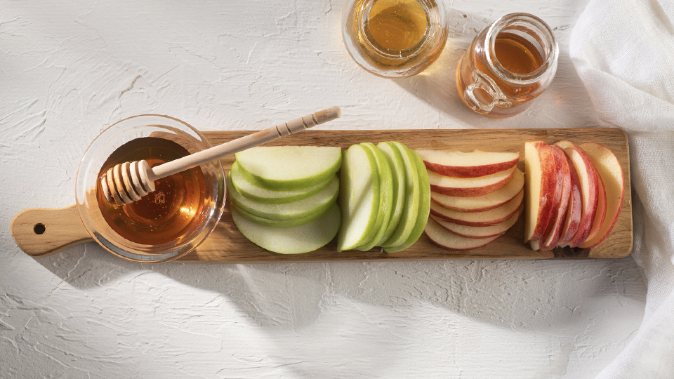 Sliced red and green apples arranged on a wooden serving board beside a bowl of honey and honey dipper.