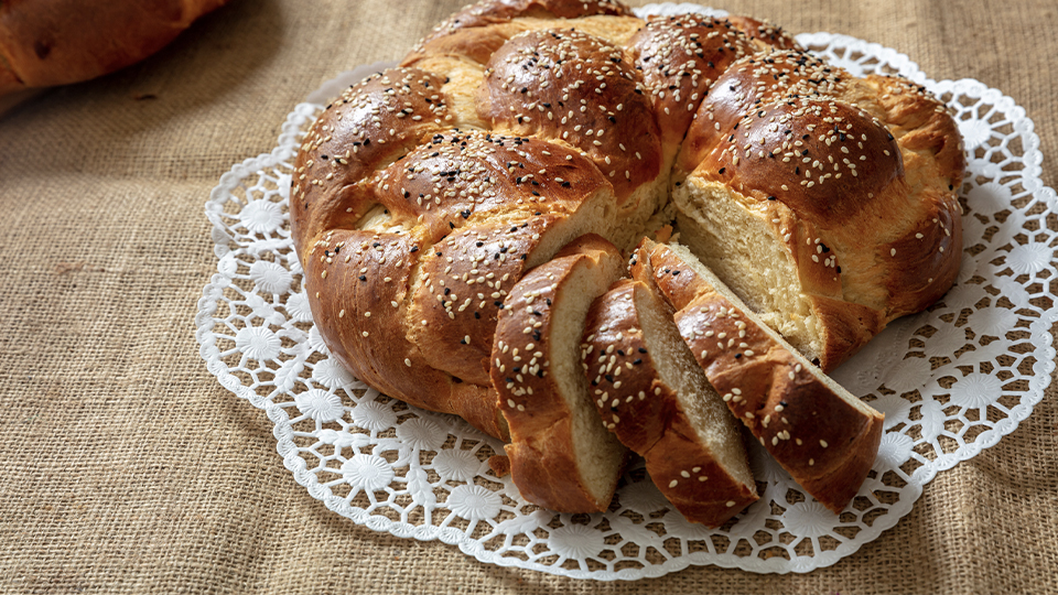 Freshly baked round challah topped with sesame seeds and partially sliced, presented on a white doily.